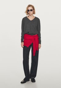 Gray v-neck sweater, red knit tied around the waist, black wide-leg jeans, and black loafers. Model wearing dark sunglasses. Plain background.
