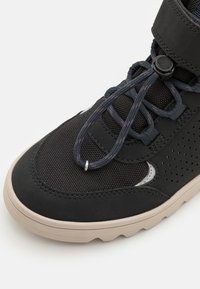 Chaussure de sport noire avec tige en mesh perforé, lacets élastiques, système de fermeture à tirette et semelle en caoutchouc beige.