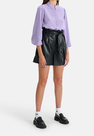 Un mannequin porte un chemisier lavande, un short noir en similicuir avec une ceinture, des chaussettes blanches et des mocassins noirs sur un fond uni.