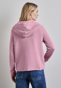 Pink textured hoodie with a hood and long sleeves, paired with blue jeans. Fabric appears soft and casual, featuring a relaxed fit design.