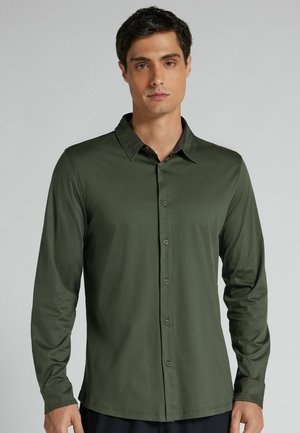 Mann trägt ein eng anliegendes, dunkelgrünes, langärmeliges Hemd mit Kragen und steht vor einem einfarbigen hellgrauen Hintergrund.