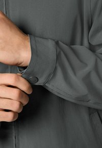 Szczegół szarej marynarki z guzikami skupia się na mankiecie o gładkiej teksturze i kontrastującym guziku. Tkanina wydaje się lekka i lekko błyszcząca.