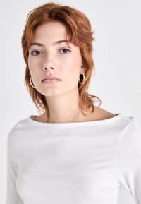 Bijela majica s dugim rukavima i brodskim dekolteom, mekanog tekstura i blago sjajna. Model nosi male srebrne obruče. Minimalistički dizajn.