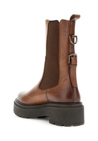 Brun läderankelboot med tjock svart sula, elastisk sidopanel, bakre dragflik och dekorativ spännband på vit bakgrund.