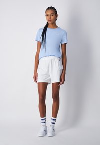 Giovane indossante una maglietta azzurra chiara, pantaloni cargo bianchi, sneakers bianche e calzini bianchi con righe blu, in piedi contro uno sfondo semplice.