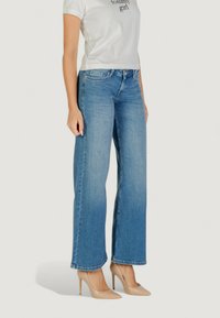 Jeans a gamba larga di un blu chiaro con una texture liscia, vita alta e orlo sfrangiato, abbinati a scarpe col tacco beige e una t-shirt bianca cropped.