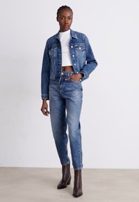 Jeans jakna s izblijedjelim plavim pranjem, ukrašena perlatnim detaljima, u kombinaciji s visokim plavim trapericama i smeškim čizmama na petu. Ispod se nosi bijeli skraćeni top.