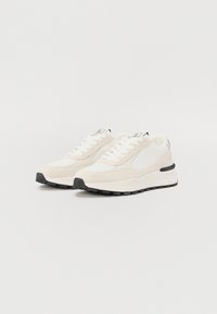 Beige semskinn og mesh sneakers med hvit såle, rund tå, lisser, svarte detaljer og en teksturert gummisåle.