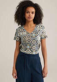 Īsu piedurkņu V-veida kakla tops ar rakstainu audumu, kurā ir zili, beigi un krēmkrāsas dizaini; apvienots ar augsto jostasvietu tumši ziliem biksēm ar sasietu jostasvietu.