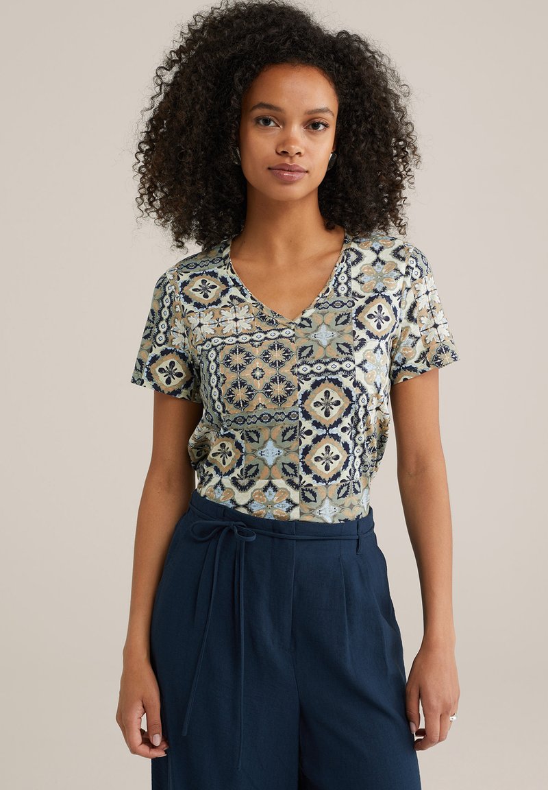 Īsu piedurkņu V-veida kakla tops ar rakstainu audumu, kurā ir zili, beigi un krēmkrāsas dizaini; apvienots ar augsto jostasvietu tumši ziliem biksēm ar sasietu jostasvietu.