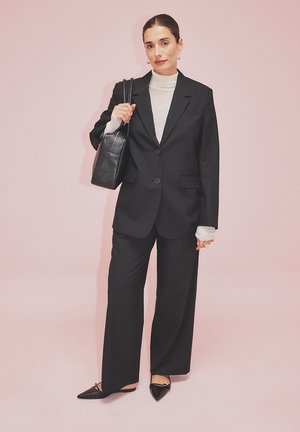 Blazer negro de gran tamaño, jersey de cuello alto blanco y pantalones de pierna ancha. Bolso negro y zapatos de punta. Fondo rosa.