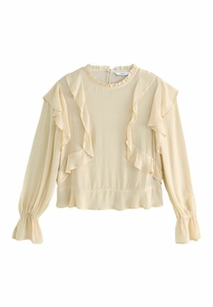Femme portant un chemisier beige à volants, un jean bleu clair avec des détails floraux subtils, et des escarpins slingback argentés à bouts pointus, debout contre un mur gris.