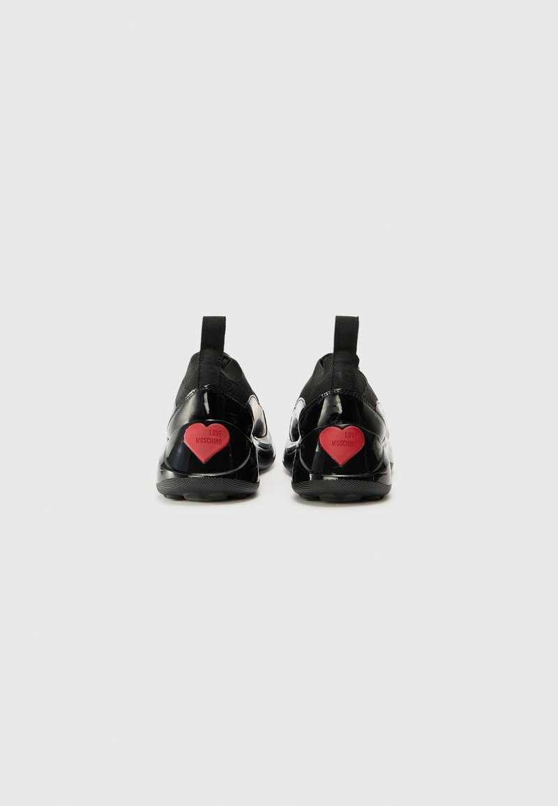 Sneakers neri con materiale lucido, forma arrotondata e un accento a forma di cuore rosso sul tallone. Presentano un'etichetta di presa e un marchio discreto.