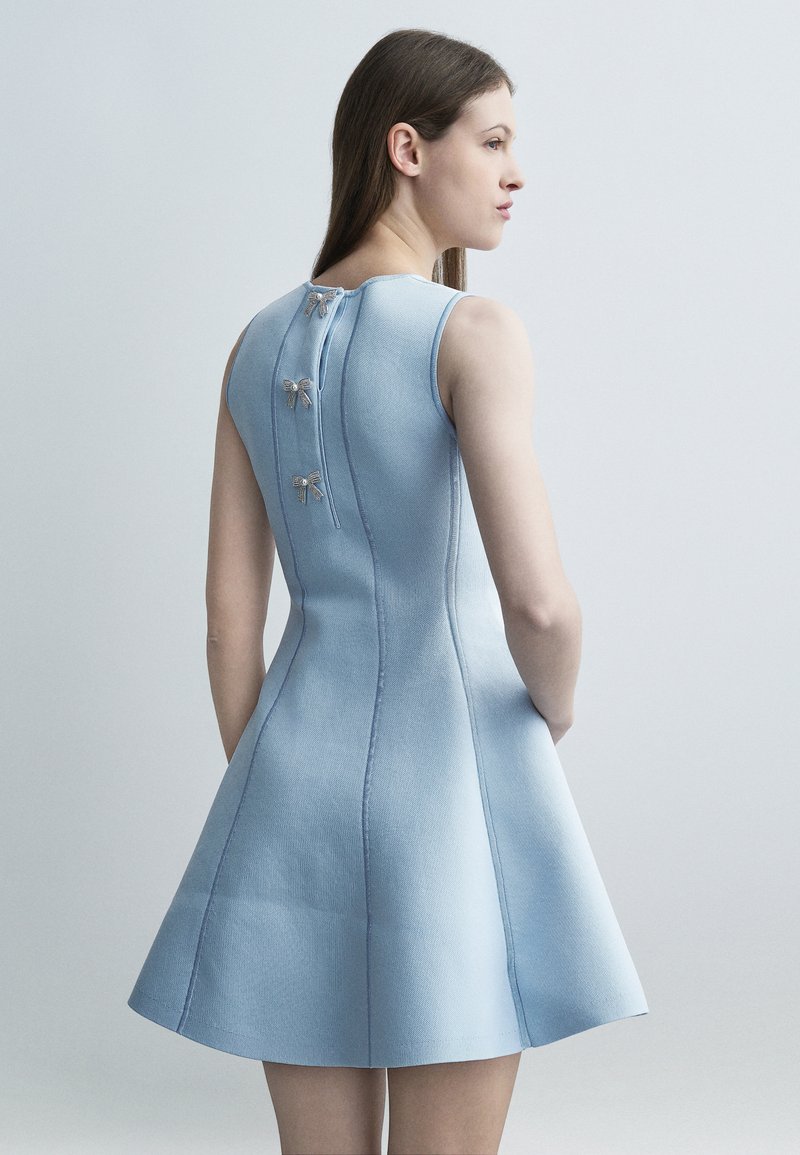 Femme portant une robe sans manches bleu clair à jupe évasée avec trois boutons décoratifs en forme de nœud sur la fermeture éclair arrière.