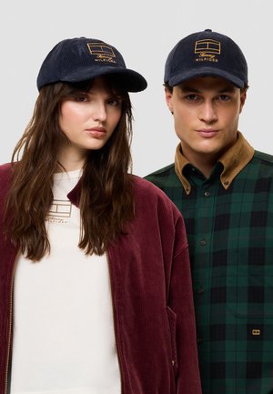 Una mujer y un hombre posan con gorras azul marino de Tommy Hilfiger; ella lleva una chaqueta burdeos y él, una camisa de cuadros verdes.
