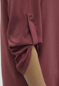 Camicia bordeaux con maniche arrotolate, tessuto morbido e dettaglio in bottone sul polsino. Esposizione della dinamica delle maniche e delle braccia.