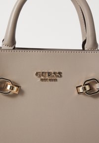 Beige håndtaske lavet af glat syntetisk materiale, med guldfarvet hardware, præget brandnavn og kæde accenter.
