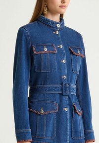 Veste en jean bleu foncé avec un col matelassé, des coutures orange, deux poches avant et une taille ceinturée. Dotée de fermetures à boutons.