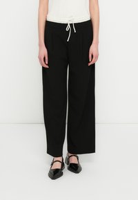 Pantalones negros de pierna ancha con cinturilla de cordón. Fabricados con tela lisa, presentan pliegues delanteros y un diseño minimalista. Combinados con zapatos negros.