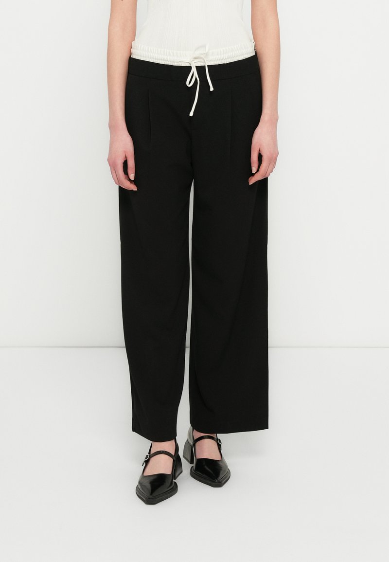 Pantalones negros de pierna ancha con cinturilla de cordón. Fabricados con tela lisa, presentan pliegues delanteros y un diseño minimalista. Combinados con zapatos negros.