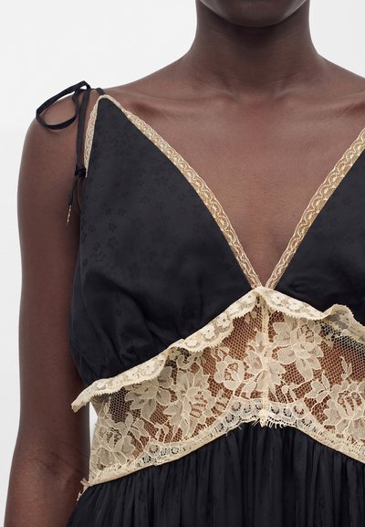 Μαύρο φόρεμα με μπεζ δαντέλα με λουλούδια γύρω από το βαθύ V λαιμό και τη μέση, λεπτές τιράντες στους ώμους σε άτομο με σκούρο δέρμα.