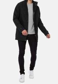 Sort vandtæt jakke med høj krave, trykknapper og lynlås, båret over en grå skjorte og mørke jeans, sammen med hvide sneakers.