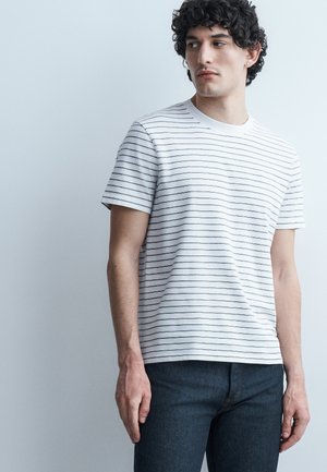 Ung mann med krøllete hår som har på seg en hvit T-skjorte med korte ermer og tynne horisontale striper, og mørke jeans, som ser mot venstre.