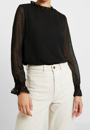 Femme portant un chemisier noir transparent à manches longues avec des pois texturés et des poignets volantés, associé à un pantalon blanc taille haute.