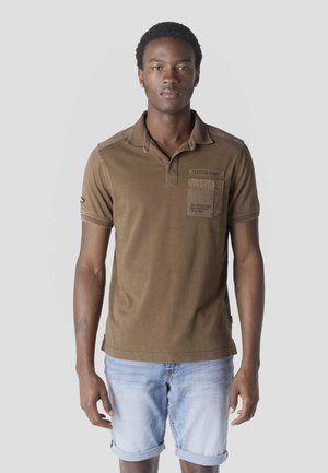 Giovane uomo che indossa una polo marrone con taschino sul petto e pantaloncini di jeans azzurri risvoltati, in piedi davanti a uno sfondo semplice.