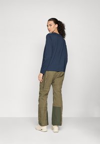 Vrouw met donker krullend haar in een marineblauw langemouwen top en olijfgroene cargobroek met versterkte boorden, staande tegen een witte achtergrond.