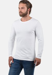 Valge pikkade varrukatega T-särk, mis on valmistatud siledast puuvillast, ümmarguse kaelusega ja istuva siluetiga, koos siniste denimist pükstega.