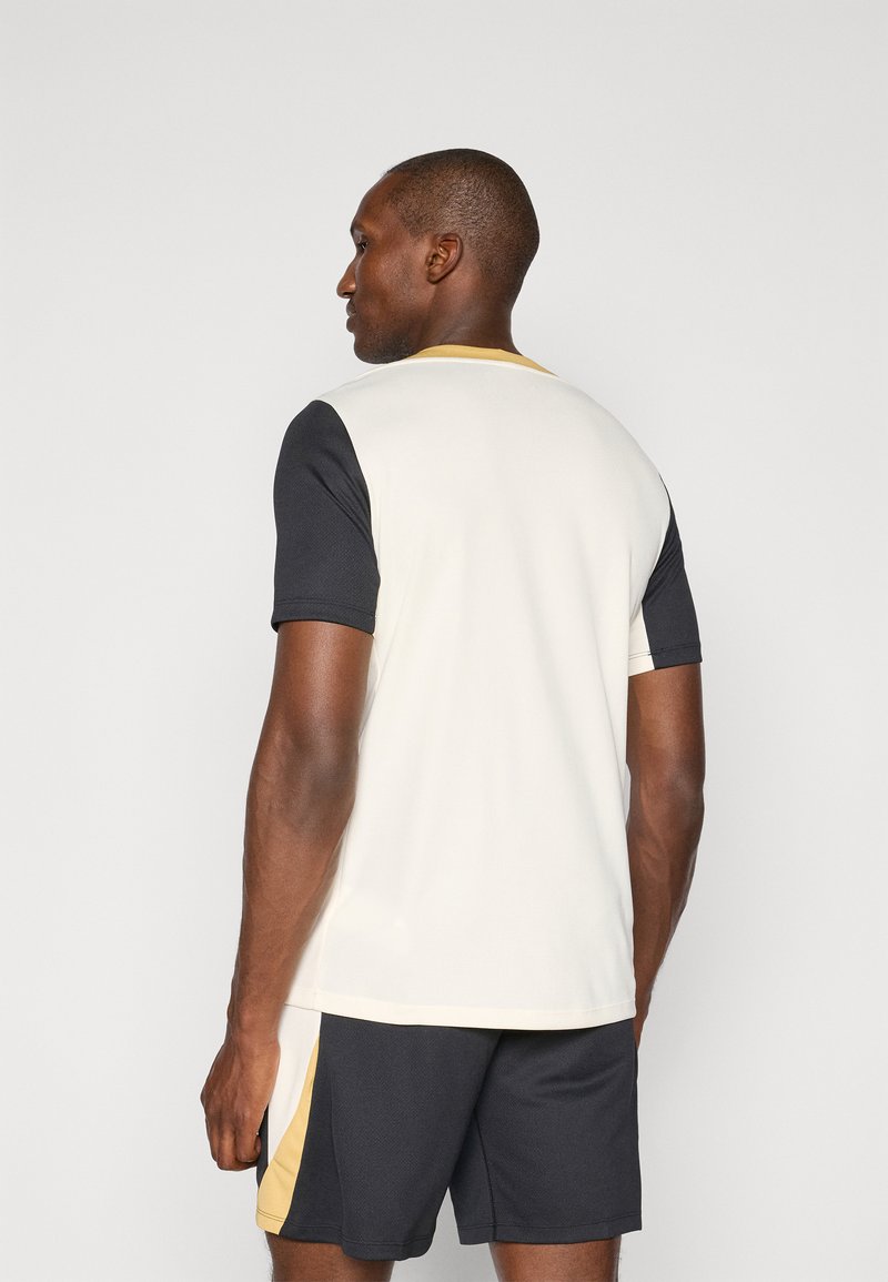 T-shirt de sport crème avec des manches noires et des accents dorés. Doté d'un col rond et d'une texture en maille. Assorti à un short noir.