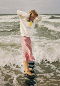 Young woman balancing barefoot on round stones in shallow ocean waves, wearing a lemon-printed sweatshirt and pink patterned skirt.