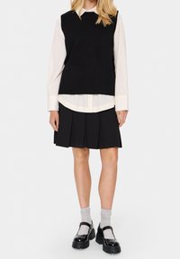 Czarny bezrękawnik nałożony na białą koszulę z kołnierzykiem, w połączeniu z czarną spódnicą plisowaną. Modelka ma na sobie szare skarpetki i czarne buty na grubej podeszwie.