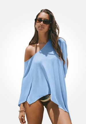 Kobieta w oversize jasnoniebieskim topie z odkrytymi ramionami, ciemnych okularach przeciwsłonecznych i kolorowych majtkach bikini na jednolitym białym tle.