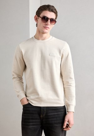 Felpa beige a girocollo con una texture sottile e un piccolo dettaglio con etichetta, abbinata a jeans scuri e indossata con occhiali da sole.