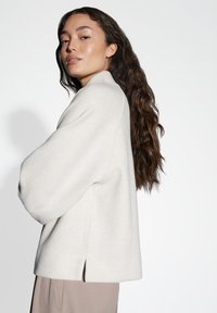 Pullover oversize grigio chiaro con colletto alto, morbido al tatto e fessure laterali, abbinato a pantaloni beige. Capelli lunghi e mossi di colore castano visibili.