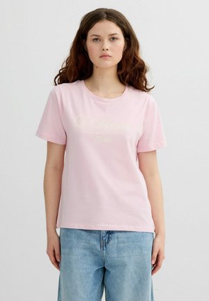 Joven con cabello castaño ondulado, vistiendo una camiseta de manga corta rosa claro y pantalones vaqueros azules, sobre un fondo liso.