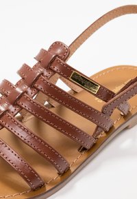 Sandal in pelle marrone con multiple cinghie orizzontali, una soletta imbottita e una placca con logo dorato. La suola è piatta e di colore tan.