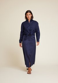 Mørkeblå denim skjortekjole med lange ermer, knappelukking foran og sidesprinkl. Har en knyttet midje-detalj og frynset kant.