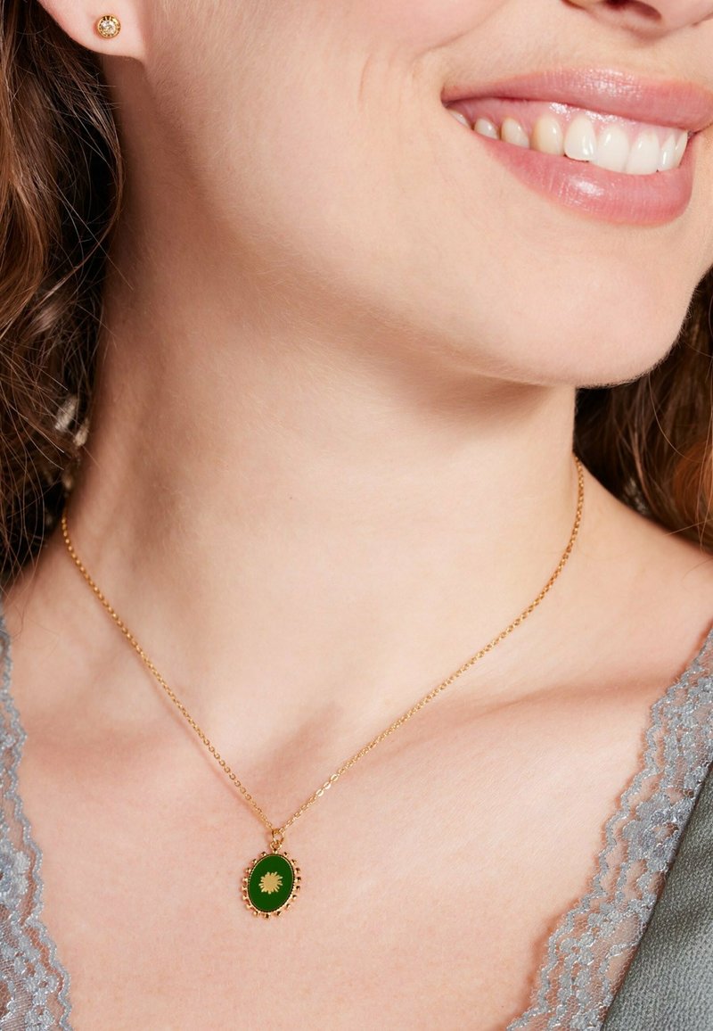 Gouden ketting met een ovale groene hanger voorzien van een bloemmotief, gedragen tegen een grijze kanttop. Eenvoudig, elegant ontwerp.