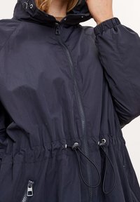 Námořnická modrá větrovka se zapínáním na zip, elastickými manžetami a stahovacími šňůrkami v pase. Disponuje bočními kapsami a kapucí. Hladká textura.