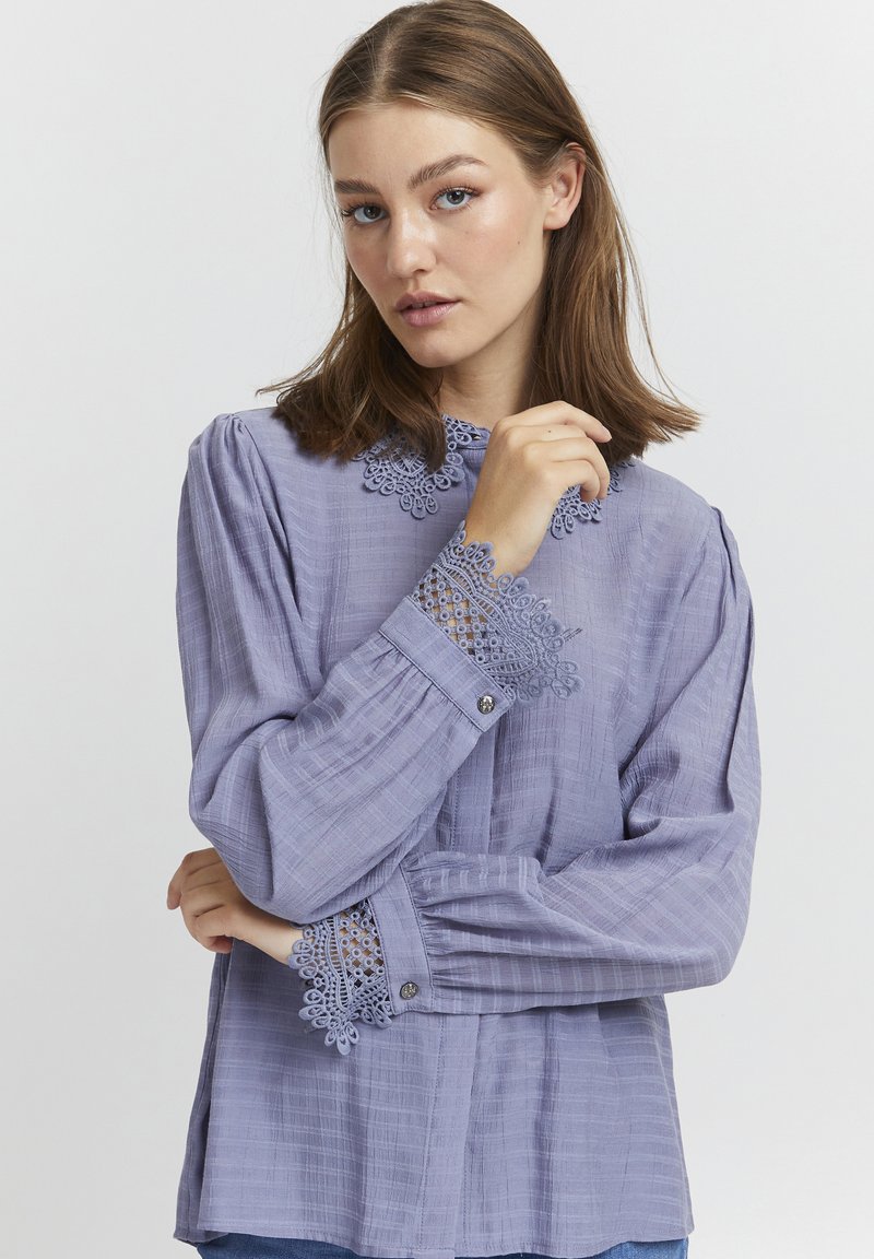 Blusa viola chiaro con maniche lunghe, tessuto testurizzato, dettagli in pizzo sui polsini e sul colletto, e chiusura frontale con bottoni.