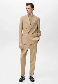 Giacca beige doppiopetto con due tasche frontali, abbinata a pantaloni affusolati. Abbinata a un maglione leggero a trama e mocassini neri.