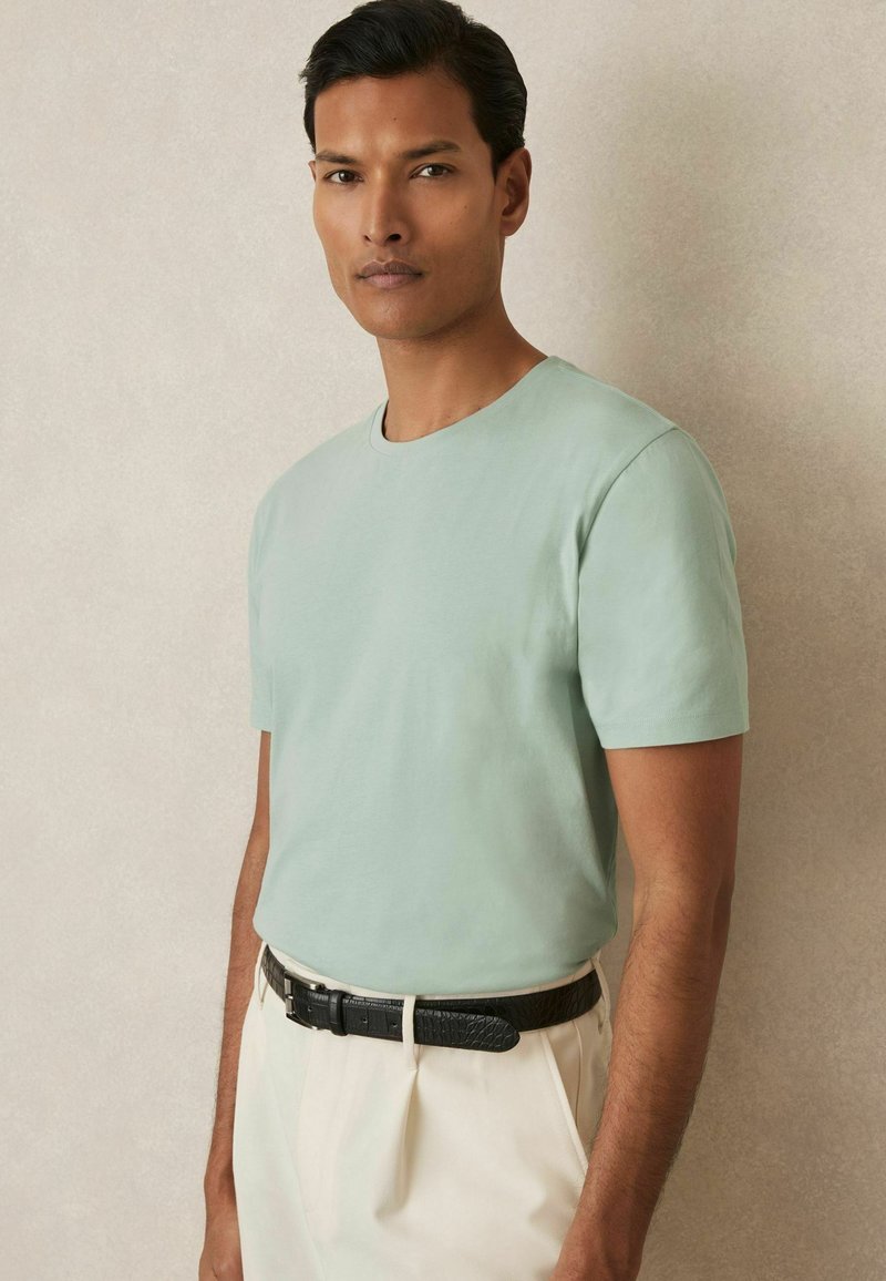 Tricou scurt de culoare verde mentă, cu guler rotund, asortat cu pantaloni crem și o curea neagră texturată, având un material din bumbac fin.