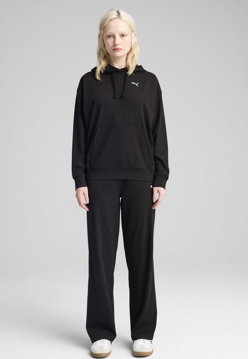 Schwarzer Hoodie mit einer Vordertasche und einer Kapuze mit Zugband, kombiniert mit passenden schwarzen Hosen; beide aus einem glatten, weichen Material gefertigt.