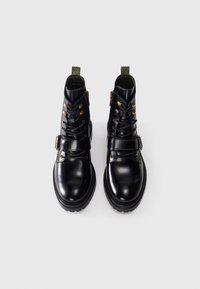 Black leather ankle boots with a glossy finish, featuring metal hardware, laces, and a strap across the midfoot. Textured sole for grip.