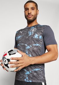 Puma short-sleeve soccer jersey in dark gray with blue abstract patterns. Player holds a black and white soccer ball with accents.