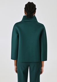 Haut surdimensionné teal avec un col montant, fabriqué à partir d'un tissu lisse et structuré. Présente des manches trois-quarts et une coupe décontractée.