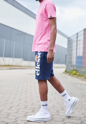 Mężczyzna stojący na zewnątrz, ubrany w różowy T-shirt, granatowe szorty Ellesse, białe skarpetki Ellesse oraz białe wysokie trampki.